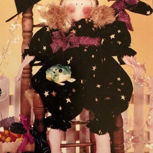 May include: A fabric doll with blonde hair, wearing a black outfit with gold stars and a purple glittery bow. The doll has rosy cheeks, a small mouth, and is holding a small green frog. A black witch hat is positioned behind the doll's head.