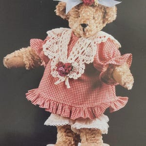 May include: A tan plush teddy bear wearing a red and white gingham dress with a white lace collar, a red fabric rose, and white lace bloomers. The bear has a light blue ribbon bow with a red rose on its head and stands on a white lace doily.