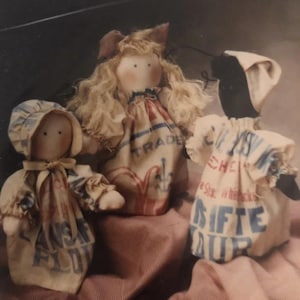 May include: Three fabric dolls with simple facial features, dressed in clothing made from vintage-style printed flour sacks. The dolls feature light and dark skin tones, with one wearing a bonnet and another with blonde yarn hair, all displaying text.