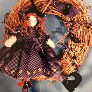 May include: A fabric doll with red yarn hair wearing a black and purple checkered dress with candy corn accents, a black witch hat, and striped stockings. The doll is positioned against a natural twig wreath with a purple bat and star garland.