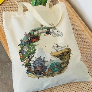 May include: A cream-coloured canvas tote bag featuring a circular, multi-coloured graphic print of various animated characters and structures, including a large castle, a dragon, and forest creatures, with two matching fabric shoulder straps.