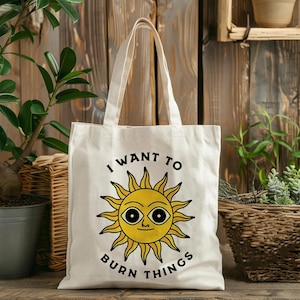 Op de afbeelding: Een witte canvas draagtas met een afbeelding van een gele zon met een gezicht en grote ogen. De tekst "I WANT TO BURN THINGS" is in zwarte hoofdletters in een boog boven en onder de illustratie van de zon op de tas gedrukt.