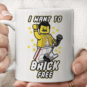 Op de afbeelding: Een witte keramische mok met een afbeelding van een geel blokfiguurtje met een snor, gekleed in een geel jasje en een witte broek, terwijl het een microfoon vasthoudt. De tekst luidt I WANT TO BRICK FREE, omringd door kleine gele sterren.