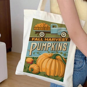 Könnte beinhalten: Eine weiße Tragetasche aus Segeltuch mit einem Grafikdruck eines hellblauen Oldtimer-Lastwagens, der orangefarbene Kürbisse auf der Ladefläche transportiert. Unter dem LKW steht der Text FALL HARVEST PUMPKINS in fetten Buchstaben über einem großen Kürbis.