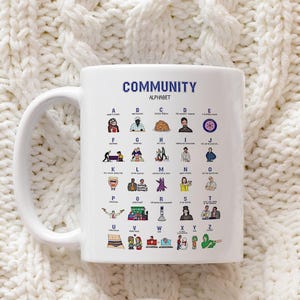 May include: A white ceramic mug featuring a blue Community Alphabet graphic. The design displays letters A through Z, each paired with a small colourful illustration and text representing characters and moments from the television show Community.