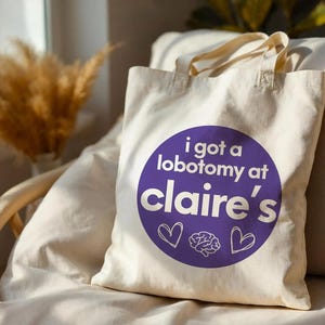 May include: A cream-colored canvas tote bag featuring a large purple circle graphic. The text reads "i got a lobotomy at claire's" in white, accompanied by two white heart outlines and a small white brain illustration centered below the text.