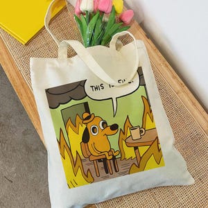 May include: A cream-colored canvas tote bag featuring a printed graphic of a cartoon dog sitting in a room filled with orange and yellow flames. The dog is wearing a hat and holding a coffee mug. A speech bubble above the dog contains the text THIS IS FINE.