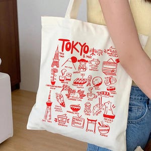 May include: A cream-colored canvas tote bag featuring a red graphic print of Tokyo landmarks, food, and cultural icons. The design includes text labels such as Tokyo Tower, Mt. Fuji, Matcha, Tsukiji Fish Market, Gyoza, and Hachiko, arranged in a grid.