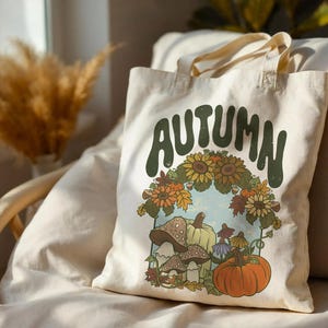 May include: A cream-colored canvas tote bag featuring a graphic print with the word AUTUMN in large green letters. The design includes a cluster of sunflowers, orange pumpkins, and brown mushrooms arranged in a decorative seasonal display.