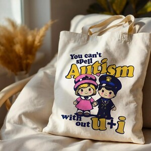 May include: A cream-colored canvas tote bag featuring a graphic print of two cartoon children, one in a pink dress and one in a dark blue uniform. The text reads, "You can't spell Autism with out u+i" in yellow and dark blue lettering.
