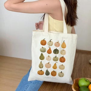 May include: A cream-colored canvas tote bag featuring a grid of twenty illustrated pumpkins and gourds in various shades of orange, green, and white. The text "pumpkin season" is printed in small, simple black letters at the bottom center.