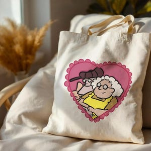 May include: A natural-colored canvas tote bag featuring a graphic print of two elderly characters inside a pink scalloped heart. One character wears a dark cap and glasses, while the other has white curly hair, glasses, and a yellow top.
