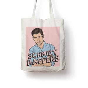 May include: A white canvas tote bag featuring a graphic illustration of a man with dark hair wearing a light blue button-down shirt. The design is set against a light pink background with the black text "SCHMIDT HAPPENS" printed in front.