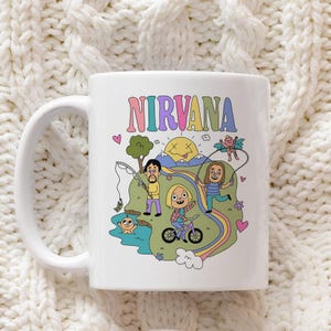 Puede incluir: Una taza de cerámica blanca con una colorida ilustración de dibujos animados y el texto NIRVANA en la parte superior. El gráfico muestra a tres personas en un paisaje con un sol, un arcoíris, un árbol, una bicicleta, una caña y un nadador.