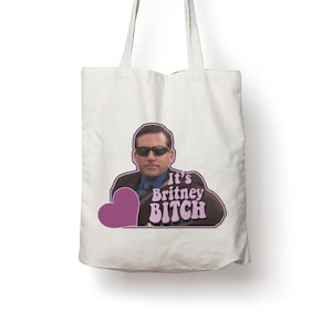 Op de afbeelding: Een witte canvas tas met een grafische print van een persoon met een donkere zonnebril en een pak. De tekst "It's Britney BITCH" staat in een lichtpaars lettertype naast een effen paarse hartvorm op de voorzijde van de tas.
