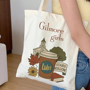 Könnte beinhalten: Eine cremefarbene Stofftasche mit einem Grafikdruck, der den Text Gilmore girls, einen Uhrturm, einen mit Luke's beschrifteten Kaffeebecher, ein rotes Ahornblatt, eine gelbe Blume, einen grünen Baum, ein rotes Buch und ein Banner mit der Aufschrift STARS HOLLOW zeigt.