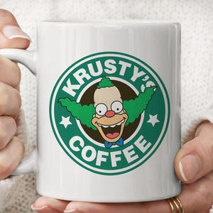 Op de afbeelding: Een witte keramische mok met een rond groen logo met de tekst KRUSTY'S COFFEE. De afbeelding toont een clownfiguur met groen haar, een rode neus en een blauwe vlinderdas, gecentreerd in de groene ring die ook twee witte sterren bevat.