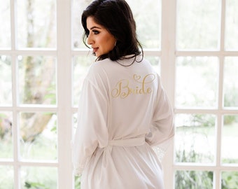 Op de afbeelding: Witte satijnen badjas met een kanten rand en het woord "Bride" in gouden letters op de rug.