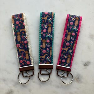 May include: Three keychains with a floral and dog print design. The keychains are in white, teal, and pink. Each keychain has a silver key ring.