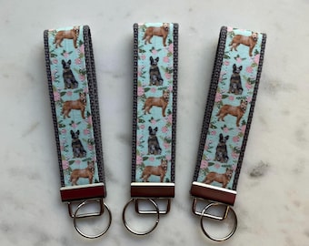May include: Three blue and white patterned keychains with silver key rings. Each keychain features a repeating pattern of Australian Shepherd dogs on a light blue background with pink flowers.
