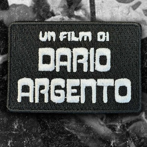 May include: A rectangular black embroidered patch featuring white stitched text that reads UN FILM DI DARIO ARGENTO. The patch has a thick black border and a textured fabric surface, with the lettering centered in a bold, sans-serif typeface.