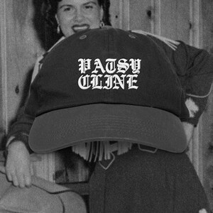 May include: A black baseball cap featuring the white embroidered text PATSY CLINE in a gothic-style font on the front panel. The cap has a curved brim and a structured crown, presented in a monochromatic black and white aesthetic.