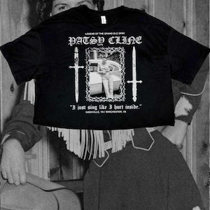 May include: A black short-sleeve t-shirt featuring a white graphic print of Patsy Cline. The design includes the text "LEGEND OF THE GRAND OLE OPRY," "PATSY CLINE," and "I just sing like I hurt inside." It also displays two swords and a barbed wire frame.