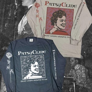 Può includere: Due maglie a maniche lunghe con un ritratto di Patsy Cline. La maglia superiore è beige con testo e grafiche rosse, quella inferiore è grigio scuro con testo e grafiche bianche. Entrambe riportano il testo Patsy Cline e Night Winds Whisper To Me.