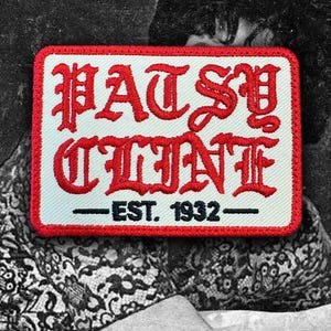 May include: A rectangular embroidered patch with a white background and a red border. The text PATSY CLINE is stitched in large red gothic-style lettering, with the black text EST. 1932 centered below it, flanked by two horizontal black lines.
