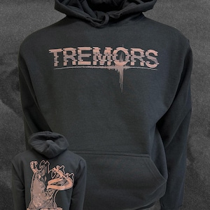 May include: A black hooded sweatshirt featuring the word TREMORS in a distressed, light-colored font on the chest. The back displays a graphic of a hand emerging from the ground with the text: THERE IS SOME HIGHER FORCE AT WORK HERE. ARE WE ASKING TOO MUCH OF LIFE?