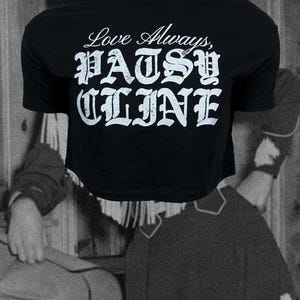 Peut inclure: Un haut court noir à manches courtes avec une inscription blanche vieillie indiquant Love Always, PATSY CLINE. Le texte est disposé avec la phrase en écriture cursive Love Always au-dessus de la police gothique grasse PATSY CLINE.