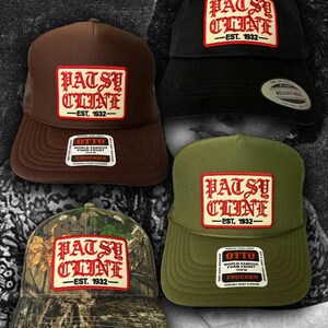 May include: Four baseball caps in brown, black, olive green, and camouflage patterns. Each cap features a rectangular patch with the red text "PATSY CLINE" and "EST. 1932" in a gothic font. The caps include various product stickers and labels.