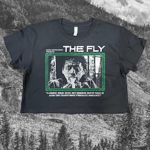 Puede incluir: Una camiseta corta negra con un estampado gráfico de la película The Fly. El diseño incluye un marco con borde verde con una imagen en blanco y negro de una persona, el texto David Cronenberg Presents The Fly y una cita sobre la película.