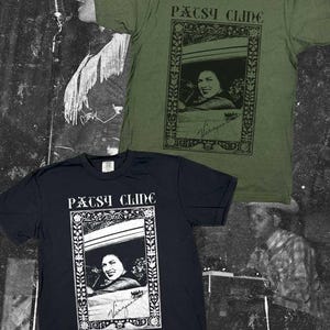 Pode incluir: Uma t-shirt verde-azeitona e uma t-shirt preta, cada uma com uma impressão gráfica de Patsy Cline. O design inclui o texto "PATSY CLINE" acima de um retrato emoldurado da cantora, com uma assinatura na parte inferior da moldura. As t-shirts têm mangas curtas.