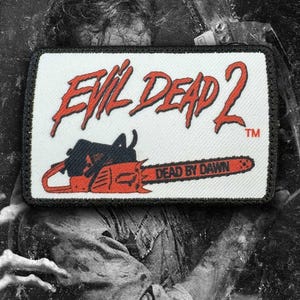 May include: A rectangular embroidered patch with a white background and a black border. The text "EVIL DEAD 2" appears in red, followed by a graphic of an orange and black chainsaw with the text "DEAD BY DAWN" printed on the blade.