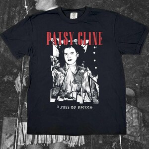 Puede incluir: Una camiseta negra de manga corta con un estampado gráfico blanco de Patsy Cline, el texto rojo "PATSY CLINE" arriba y el texto blanco "I FALL TO PIECES" abajo. El diseño incluye un retrato estilizado y fragmentado de la cantante.