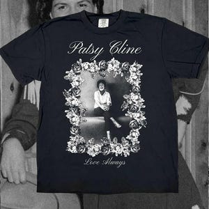 May include: A black short-sleeve t-shirt featuring a white graphic print of Patsy Cline. The design includes a rectangular floral border surrounding a portrait of the artist, with the text Patsy Cline above and Love Always printed below.