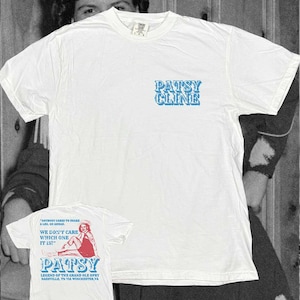 May include: A white short-sleeve t-shirt featuring the blue text PATSY CLINE on the front chest. The back displays a graphic of a person sitting, with the text ANYBODY CARES TO SHAKE A LEG, GO AHEAD. WE DON'T CARE WHICH ONE IT IS! PATSY LEGEND OF THE GRAND OLE OPRY NASHVILLE, TN VIA WINCHESTER, VA.