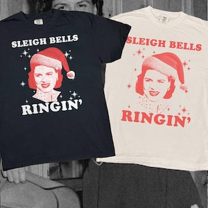 Puede incluir: Dos camisetas de manga corta, una negra y otra blanca, cada una con el gráfico de una persona con gorro de Papá Noel y el texto "SLEIGH BELLS RINGIN'" en fuente negrita. El diseño incluye pequeños detalles de estrellas alrededor del retrato.