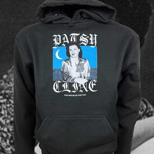 Peut inclure: Un sweat-shirt à capuche noir avec une impression graphique de Patsy Cline. Le motif comprend un fond rectangulaire bleu avec un croissant de lune, le texte PATSY CLINE en police gothique et la phrase FEELING BLUE FOR YOU sous l'image.