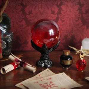 Puede incluir: Una bola de cristal roja sobre un soporte negro con forma de garra, con un fondo oscuro. Junto a ella, dos pergaminos enrollados atados con cinta roja, una botella negra con adornos dorados y una pequeña botella roja. Tema místico.