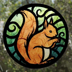 Op de afbeelding: Een ronde zonnevanger met een gestileerde oranje eekhoorn met een grote, pluizige staart. De eekhoorn is geplaatst tegen een achtergrond van groene en blauwgroene getextureerde panelen, omsloten door een stevig zwart frame met krullende details.