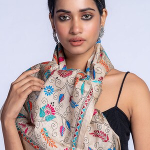 May include: A beige silk scarf with colorful floral embroidery. The scarf is draped around the neck of a person wearing a black tank top.
