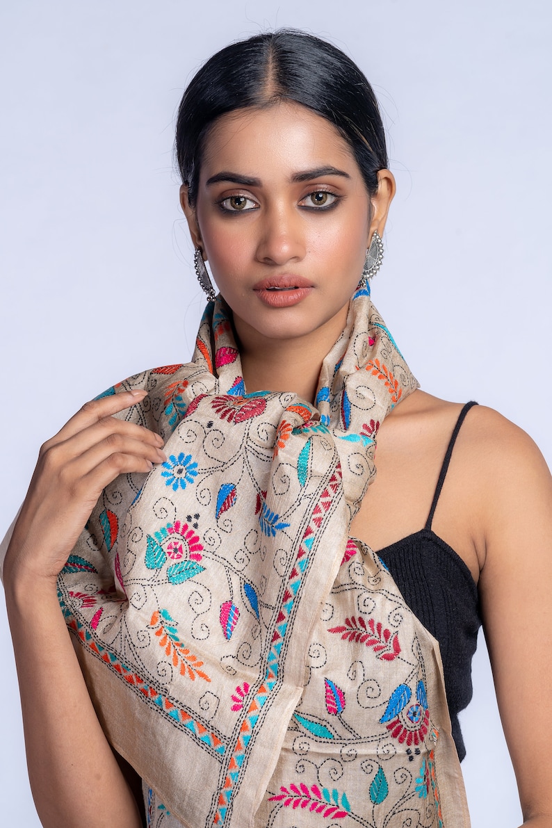 May include: A beige silk scarf with colorful floral embroidery. The scarf is draped around the neck of a person wearing a black tank top.