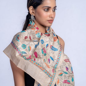 May include: A beige silk scarf with colorful floral embroidery. The scarf is folded and draped around a person's neck.