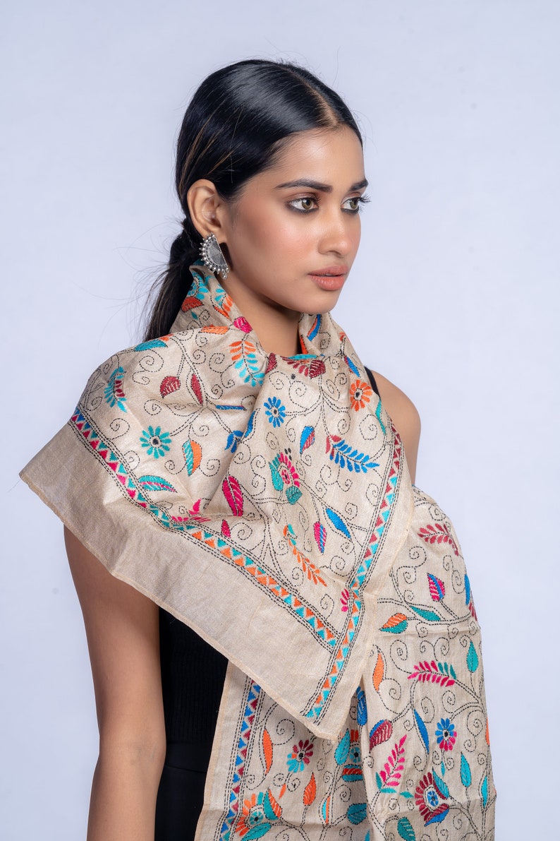 May include: A beige silk scarf with colorful floral embroidery. The scarf is folded and draped around a person's neck.