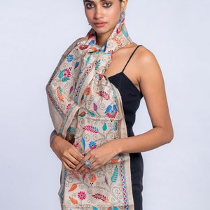 May include: A beige silk scarf with colorful floral embroidery. The scarf is tied around the neck of a person wearing a black tank top.
