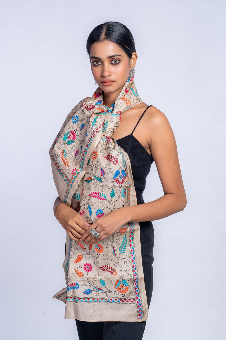 May include: A beige silk scarf with colorful floral embroidery. The scarf is tied around the neck of a person wearing a black tank top.