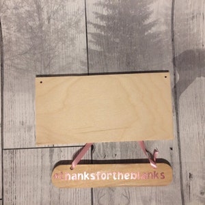 May include: A wooden sign with the text "#thanksfortheblanks" in pink lettering. The sign is hanging from two pink ribbons.