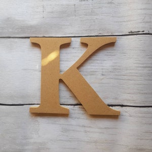 May include: A wooden letter K, painted in a light brown colour. The letter is standing on a white wooden surface.